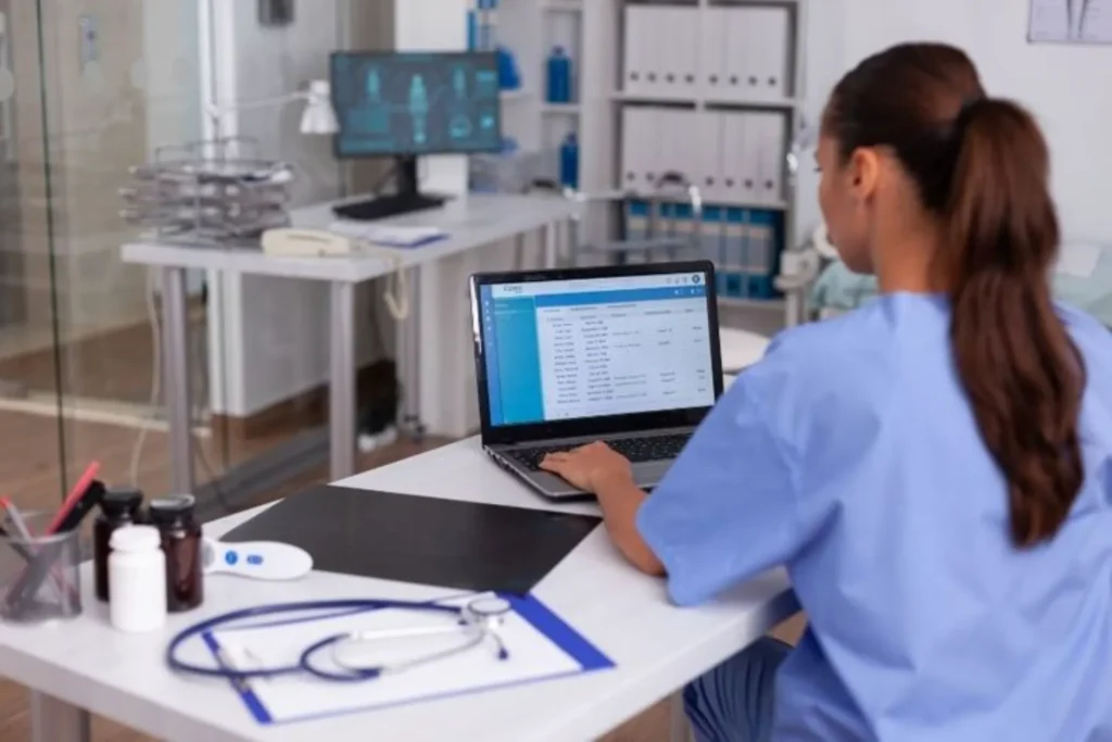 Infirmière en blouse bleue utilisant un logiciel médical sur ordinateur portable dans un cabinet.