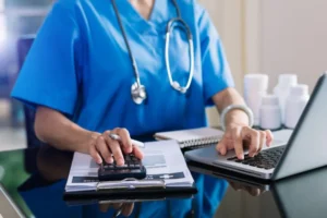 Infirmière en blouse bleue utilisant un ordinateur et une calculatrice pour gérer sa comptabilité.
